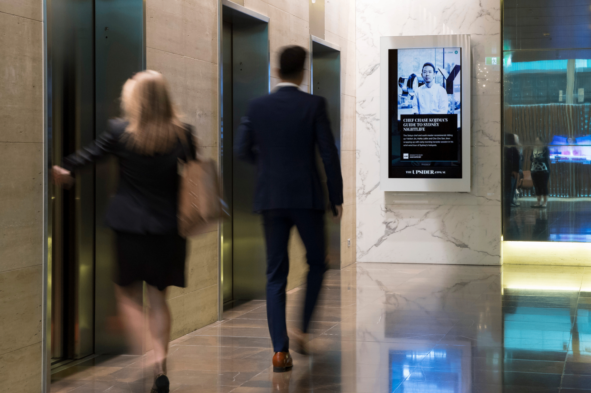 In-building digital screen in a marble office tower lobby beside the elevator banks