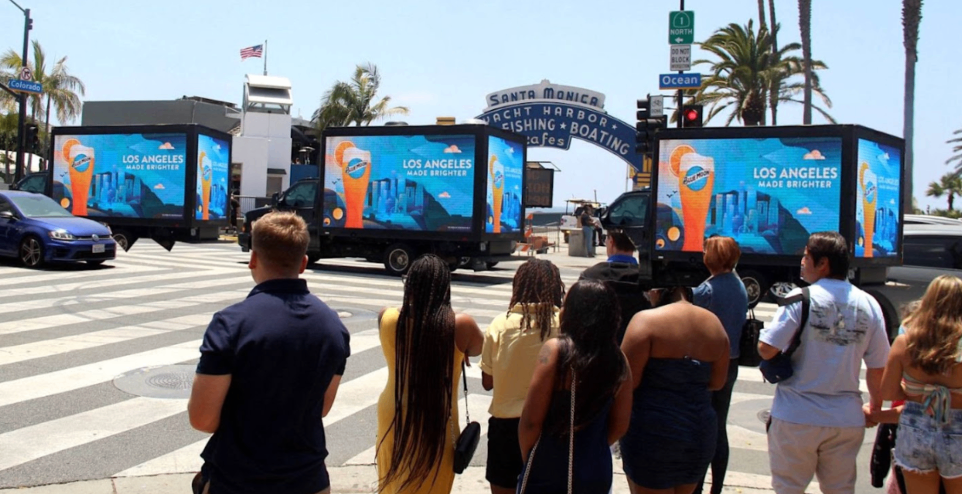 Three LED billboard trucks parked in formation outside the Santa Monica Pier, drawing a crowd of pedestrians.