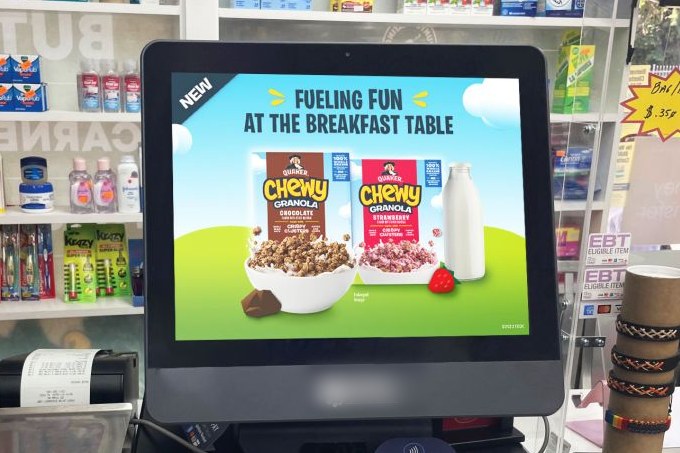 A point-of-sale screen at a convenience store checkout counter running a breakfast cereal ad, with snack and beverage shelving behind it.