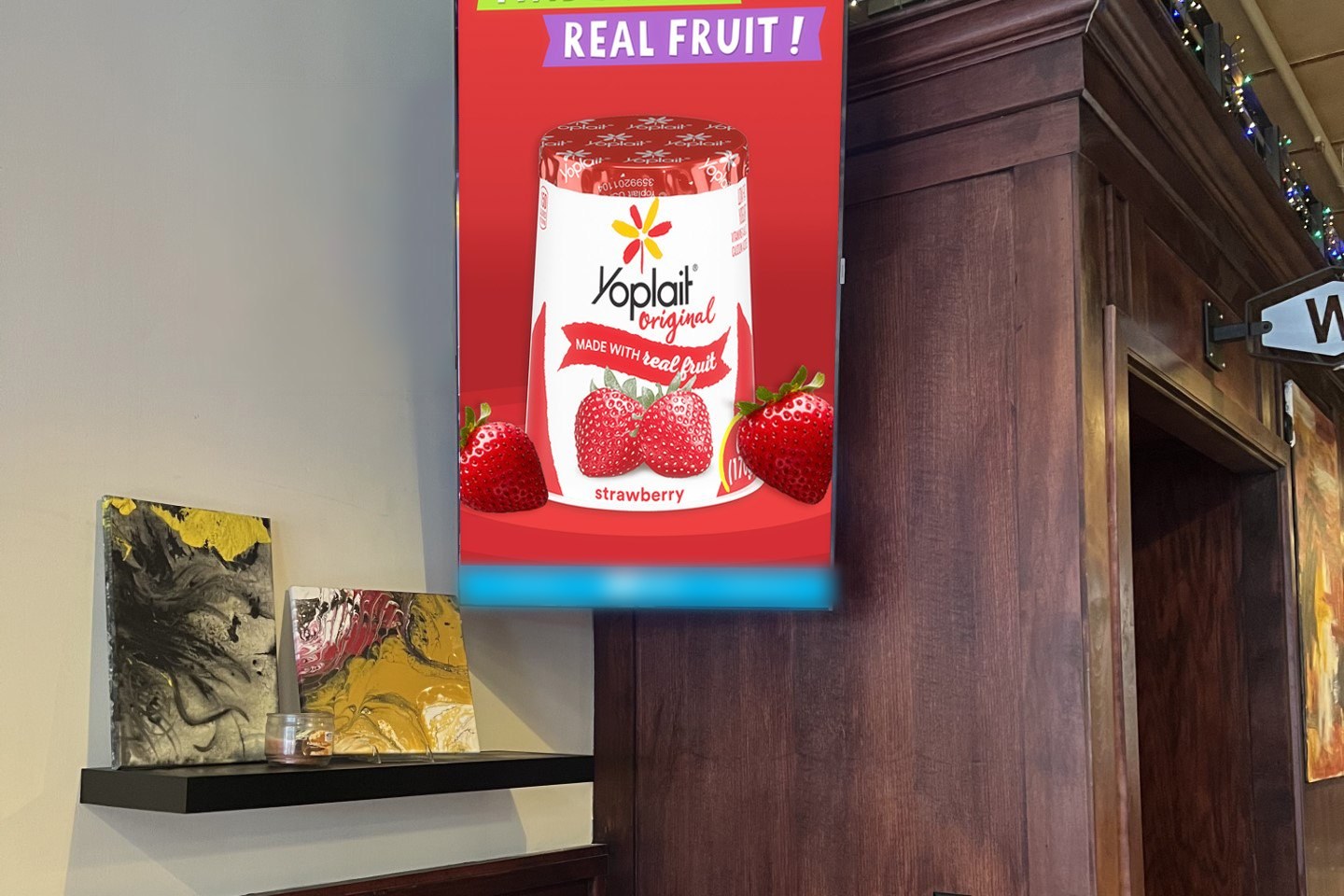 A vertical digital screen mounted on a restaurant wall running a Yoplait Original strawberry yogurt ad reading Real Fruit, beside artwork on a shelf.