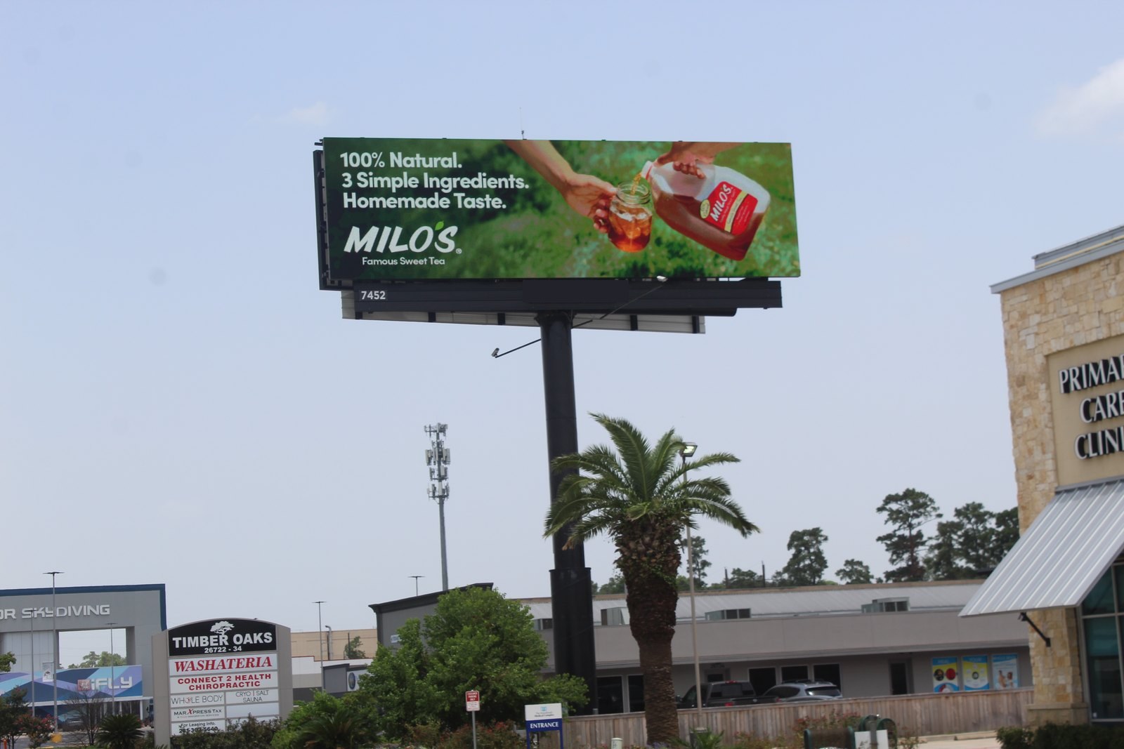 A digital billboard in a retail plaza running an ad for a sweet tea brand reading 100% Natural, 3 Simple Ingredients, Homemade Taste, with storefronts and palm trees behind it.