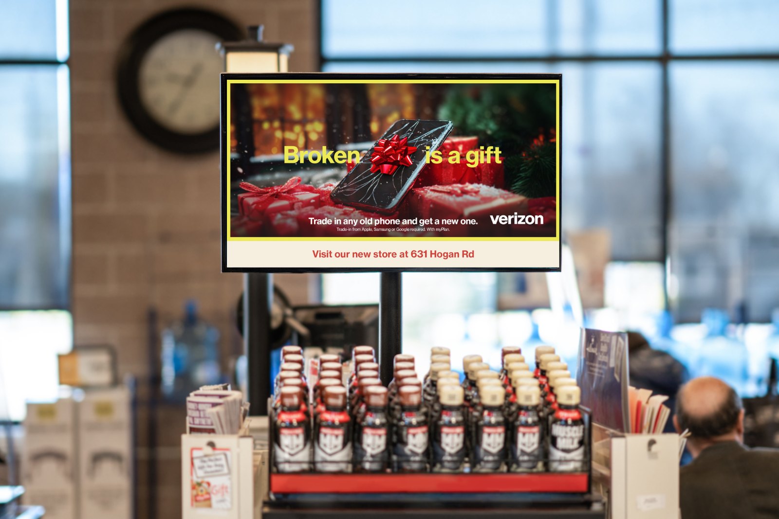 A digital screen mounted above a grocery store end-cap display running a Verizon trade-in ad, with bottled product stacked on the shelving beneath it and shoppers in the background.