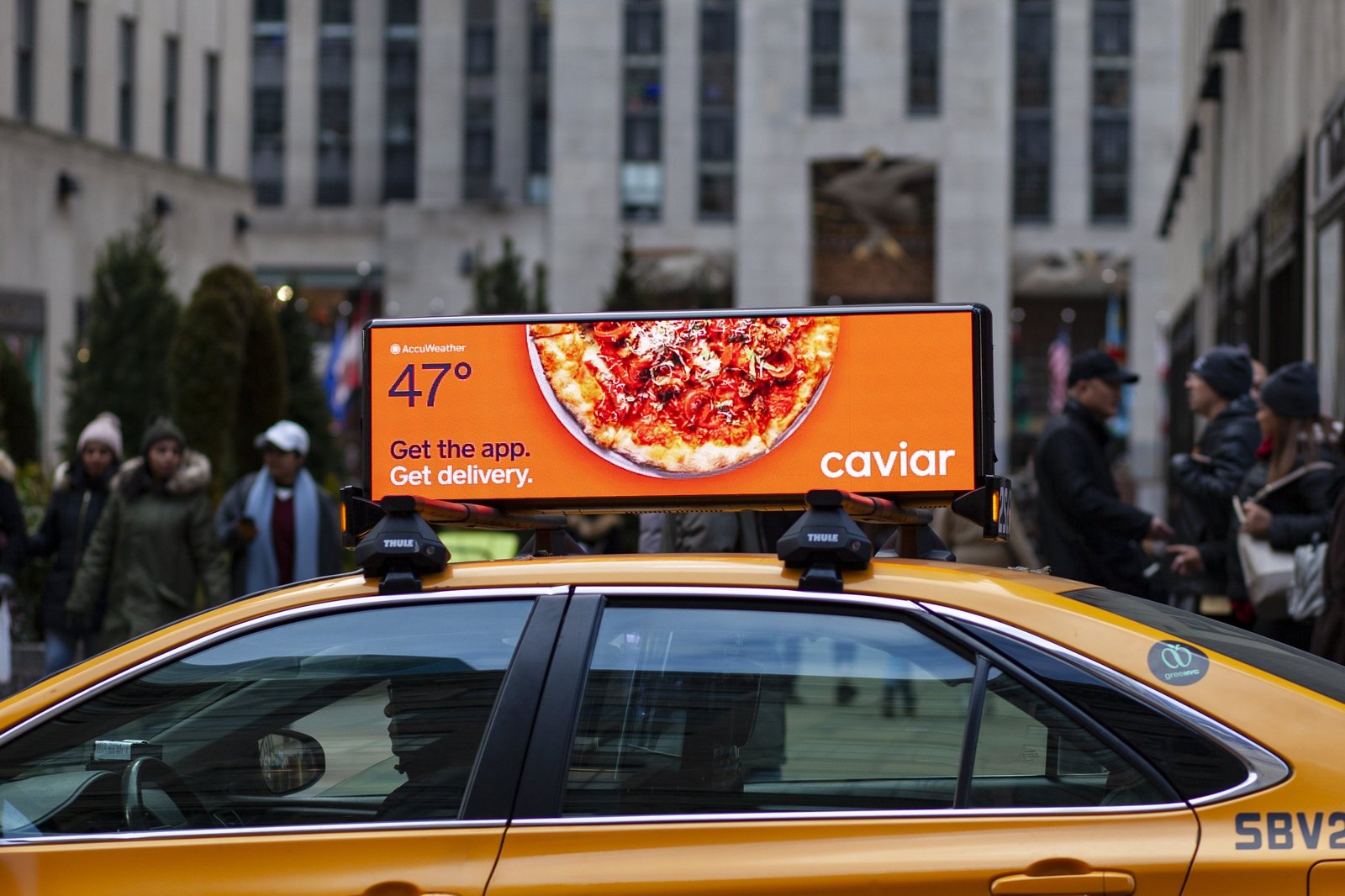 A digital taxi-top display on a yellow cab in a city street running a food-delivery ad showing a pizza, with pedestrians on the sidewalk behind.