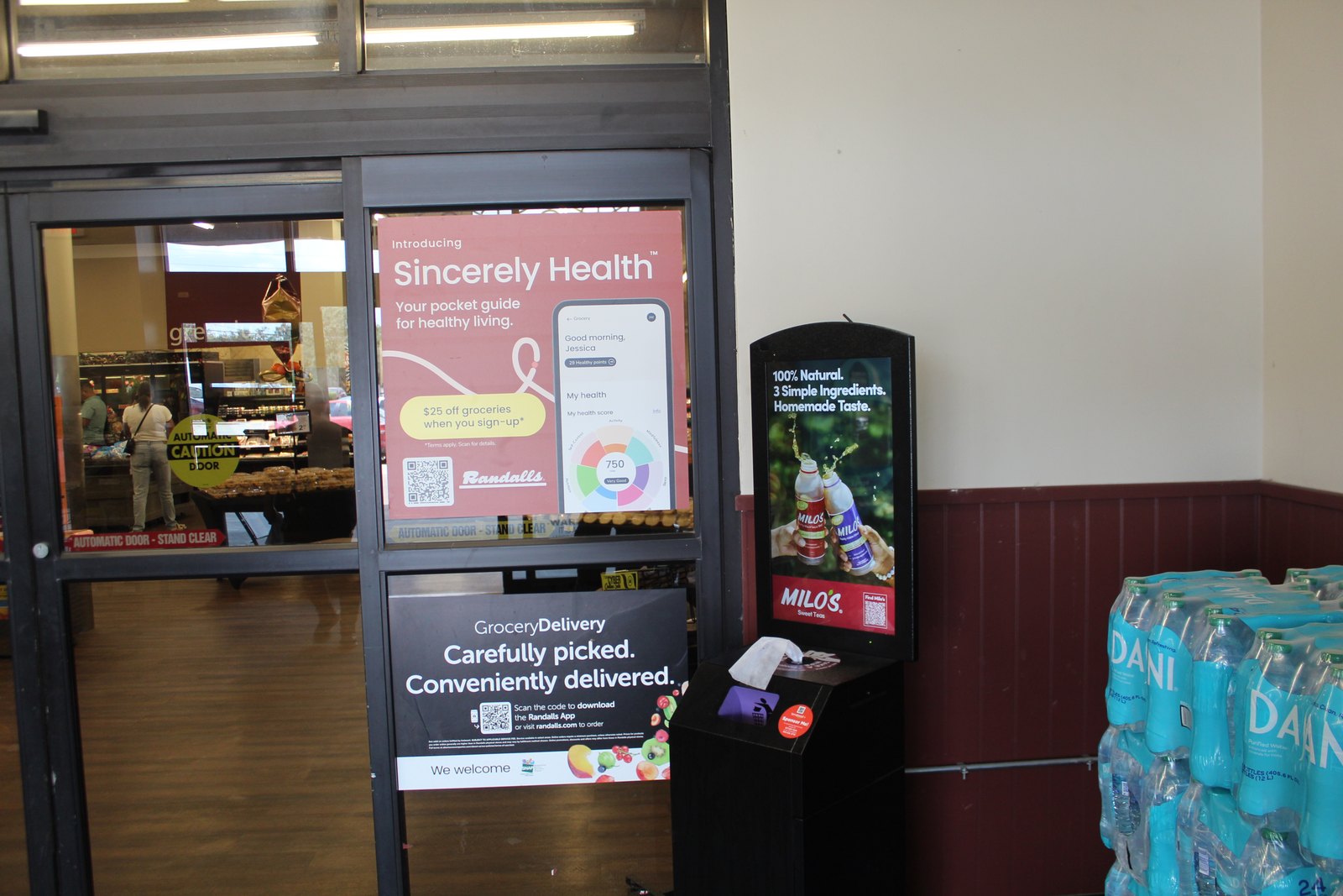 Two digital screens mounted at a grocery store entrance, one running a health guide and one running an ad for a sweet tea brand, beside stacked cases of bottled water.