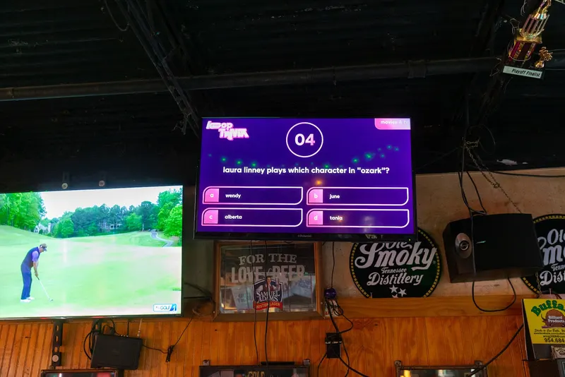 Screens over the bar at a sports bar, one showing a live broadcast and one running bar trivia.